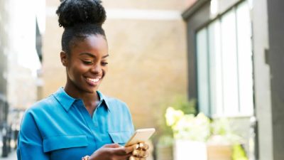 A woman standing in a blue shirt smiles as she uses her mobile phone to text message someone