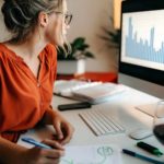 A woman studying share market stats on a computer while writing a report.