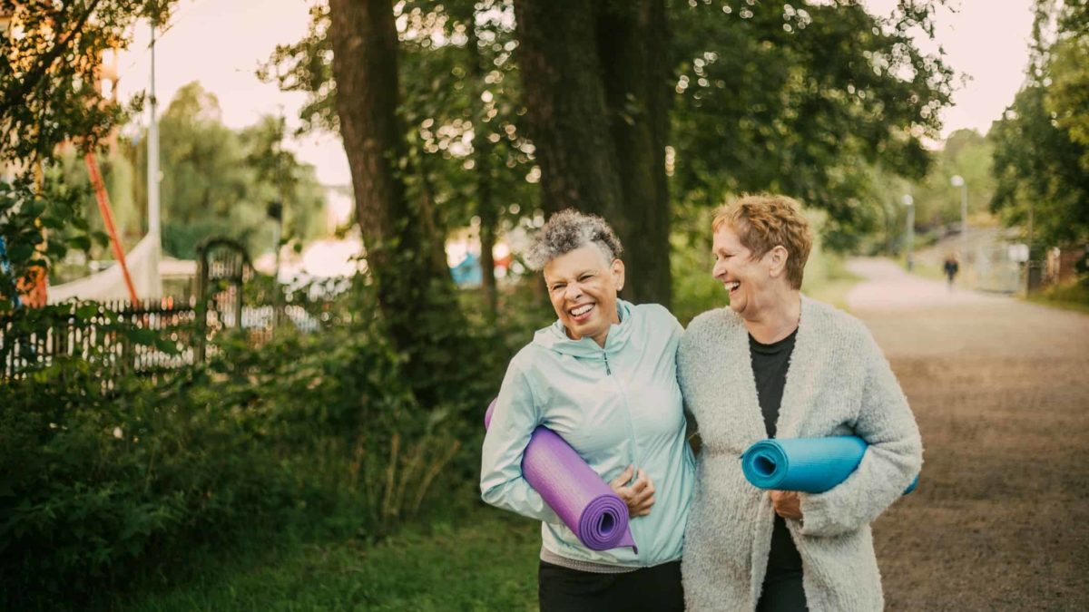 Two older women with yoga mats laughing and walking.