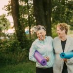 Two older women with yoga mats laughing and walking.