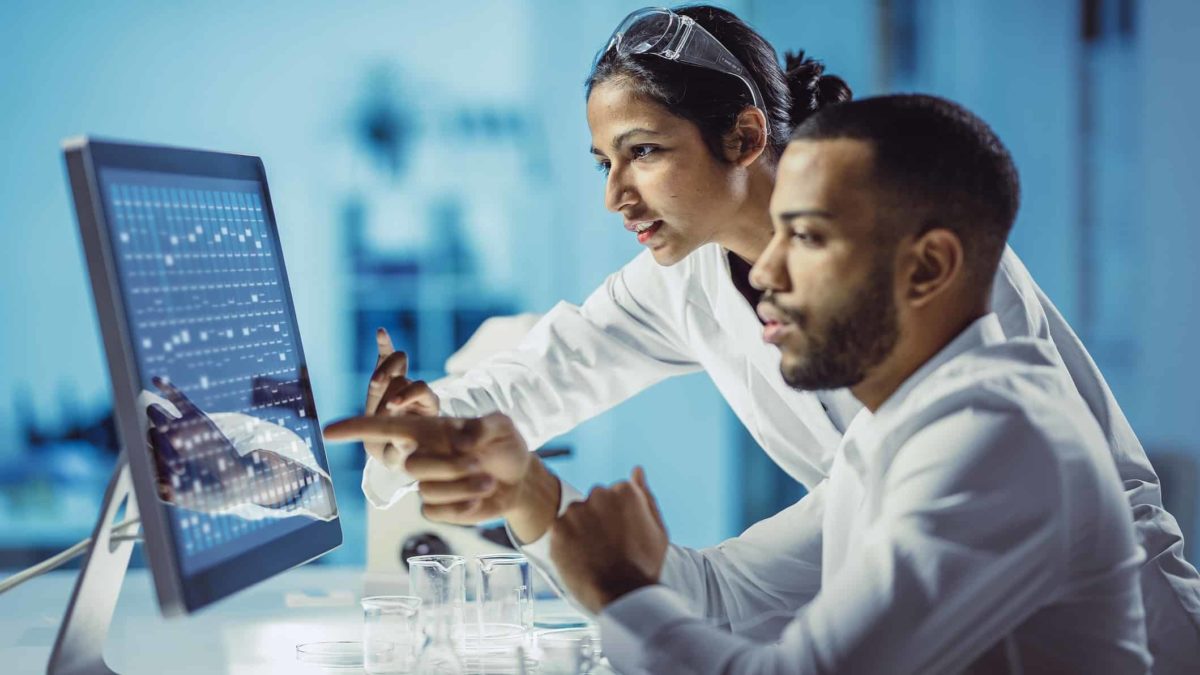 Scientists in a laboratory look at a computer screen with anticipation on their faces representing a potential change in the performance of ASX biotech shares in FY23