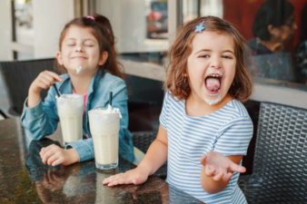 Two young girls drinking milkshakes with milk around their mouths.