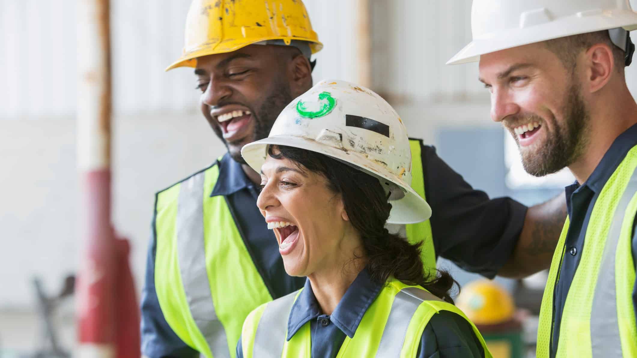 three builders in hard hats on work site looking happy