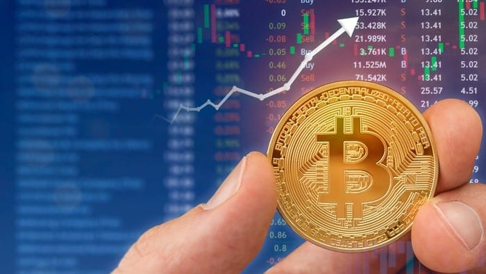 A photo of a blue graphic in the background with a white upwards arrow and a gold bitcoin being held in front of it