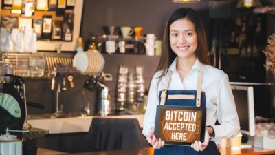 A young woman working in a coffee shop holds a sign saying: 'Bitcoin accepted here'
