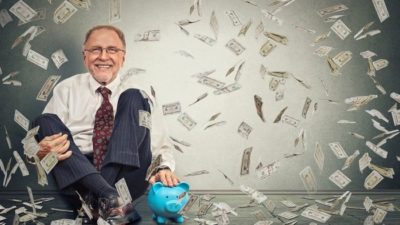 An older executive man dressed in suit trousers and a white shirt sits against a wall smiling with cash rains down over him representing dividend shares like BHP, FMG and Newcrest paying dividends in retirement