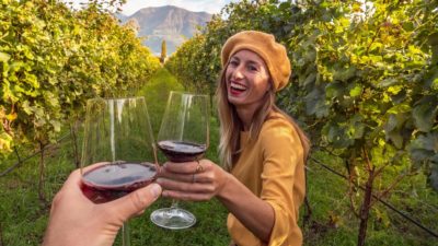 Happy smiling young woman drinking red wine while standing among the grapevines in a vineyard.