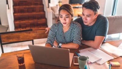 A man and woman sit at a desk staring intently at a laptop screen with papers next to them.