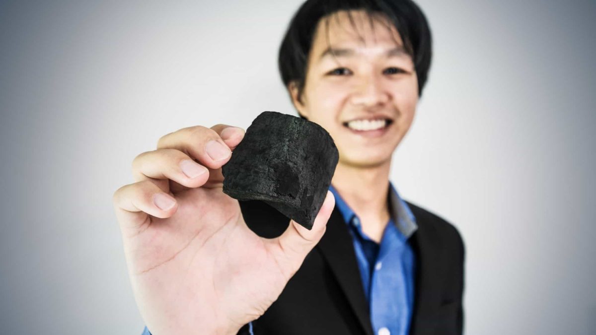 A smiling man wearing a collared blue shirt and black jacket holds a piece of black rock containing rare earths being sold by Australian Strategic Materials
