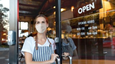 Cafe working wearing face mask and apron opening door with Open sign in window.