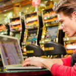 Young man sitting at a table in front of a row of pokie machines staring intently at a laptop. looking at the Crown Resorts share price