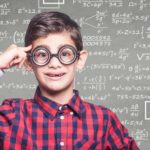 School boy wearing glasses standing in front of chalk board with maths and share price calculations on it.