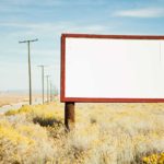 An advertising billboard with no message at the side of a lonely road in the countryside with weeds on the ground and a blue sky.