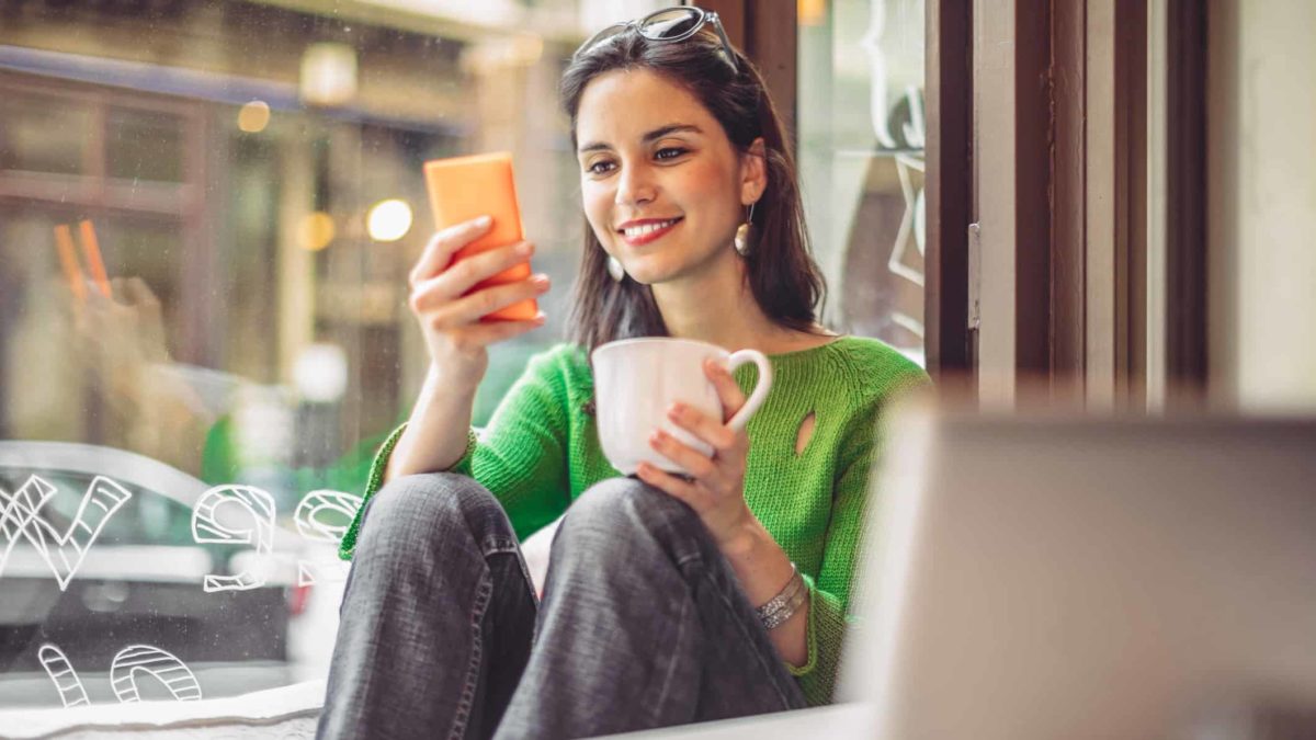 A smiling woman sits in a cafe reading a story on her phone about Rio Tinto and drinking a coffee with a laptop open in front of her.