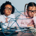 Two children sit amid a tangle of wires at a desk looking sad and despondent.