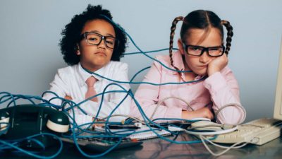 Two children sit amid a tangle of wires at a desk looking sad and despondent.