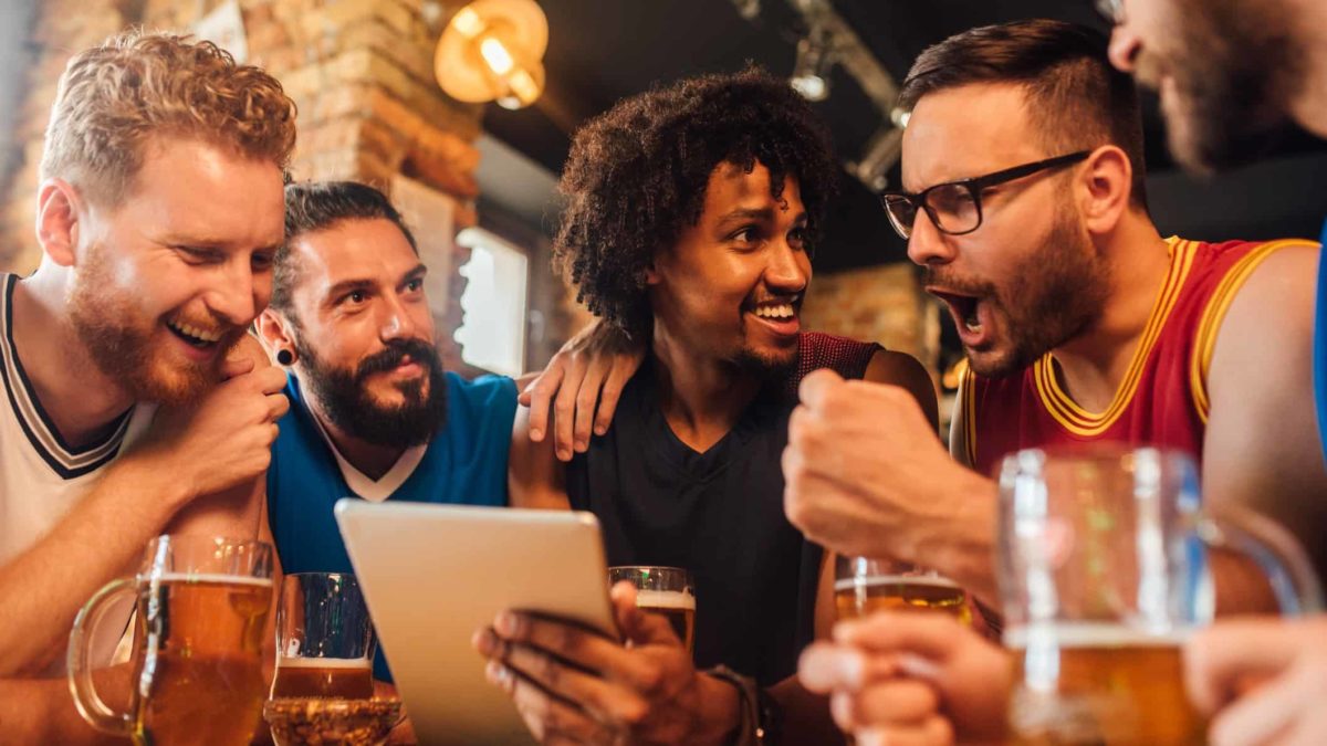 a group of men sitting together at a bar looking over an online device and celebrating.