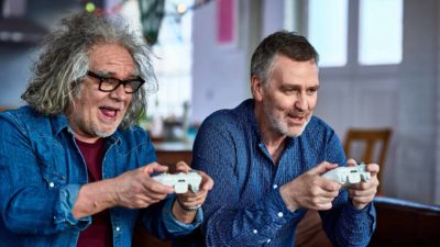 Two guys, one middle aged one older, play a computer game intently but with smiles on the couch.