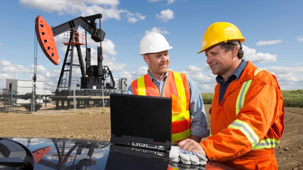 Two miners wearing hard hats standing at a mining site in front of a laptop computer