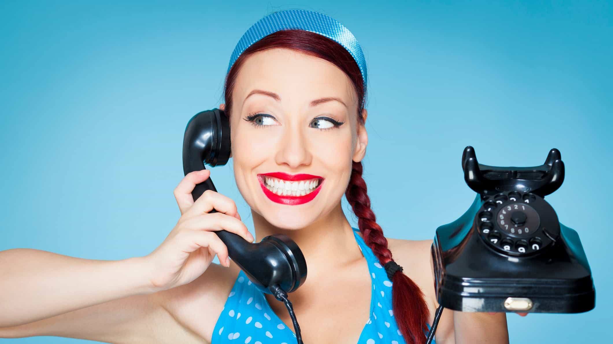 A woman smiles widely while using an old fashioned hand set telephone with dial.