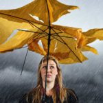 A woman with a sad face stands under a shredded umbrella in a grey thunderstorm.