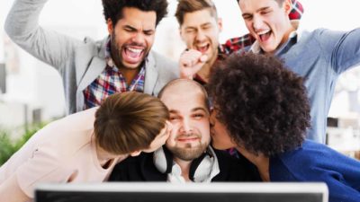 A group of people crowd around a guy sitting at a computer, and two plant kisses on his cheeks, the rest cheering.