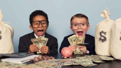two children dressed in business attire with joyous, wide-mouthed expressions count money at a desk covered in cash and sacks of money either side.