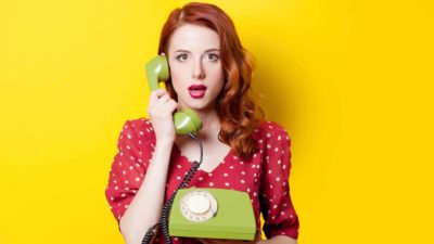 A young woman in a red polka-dot dress holds an old-fashioned green telephone set in one hand and raises the phone to her ear representing the Telstra share price and the opportunity for investors in FY23