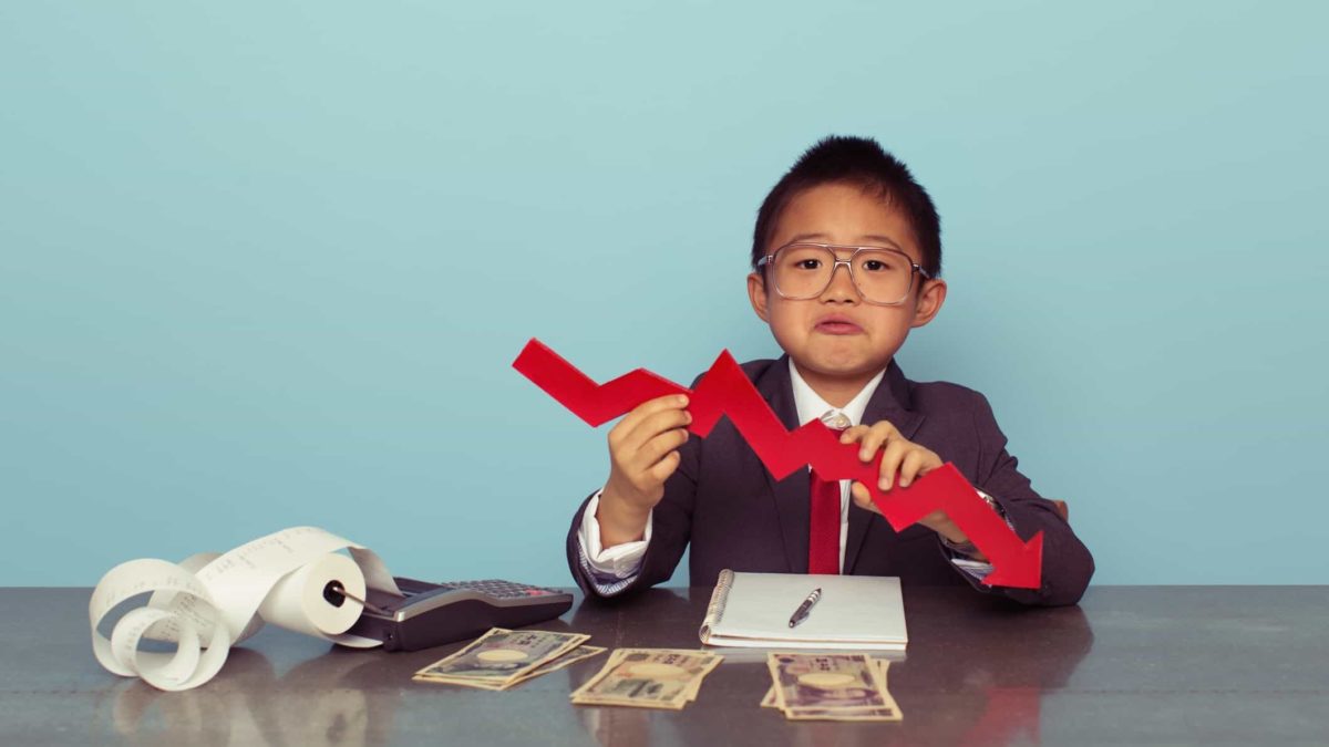 A child in full business suit holds a falling, zigzagged red arrow pointing downwards while sitting at a desk that holds cash and an old-fashioned adding machine with paper spooling.