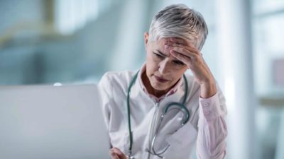 a doctor with stethoscope around neck sits as a computer with head in hand, looking despondent.