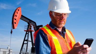 oil and gas worker checks phone on site in front of oil and gas equipment