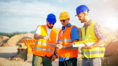 Three miners stand together at a mine site studying documents with equipment in the background