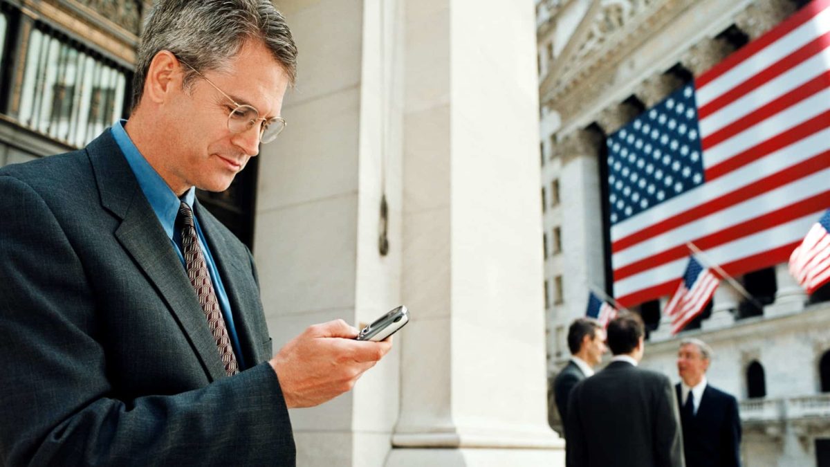 a business person checks his mobile phone outside a Wall Street office with an American flag and other business people in the background.
