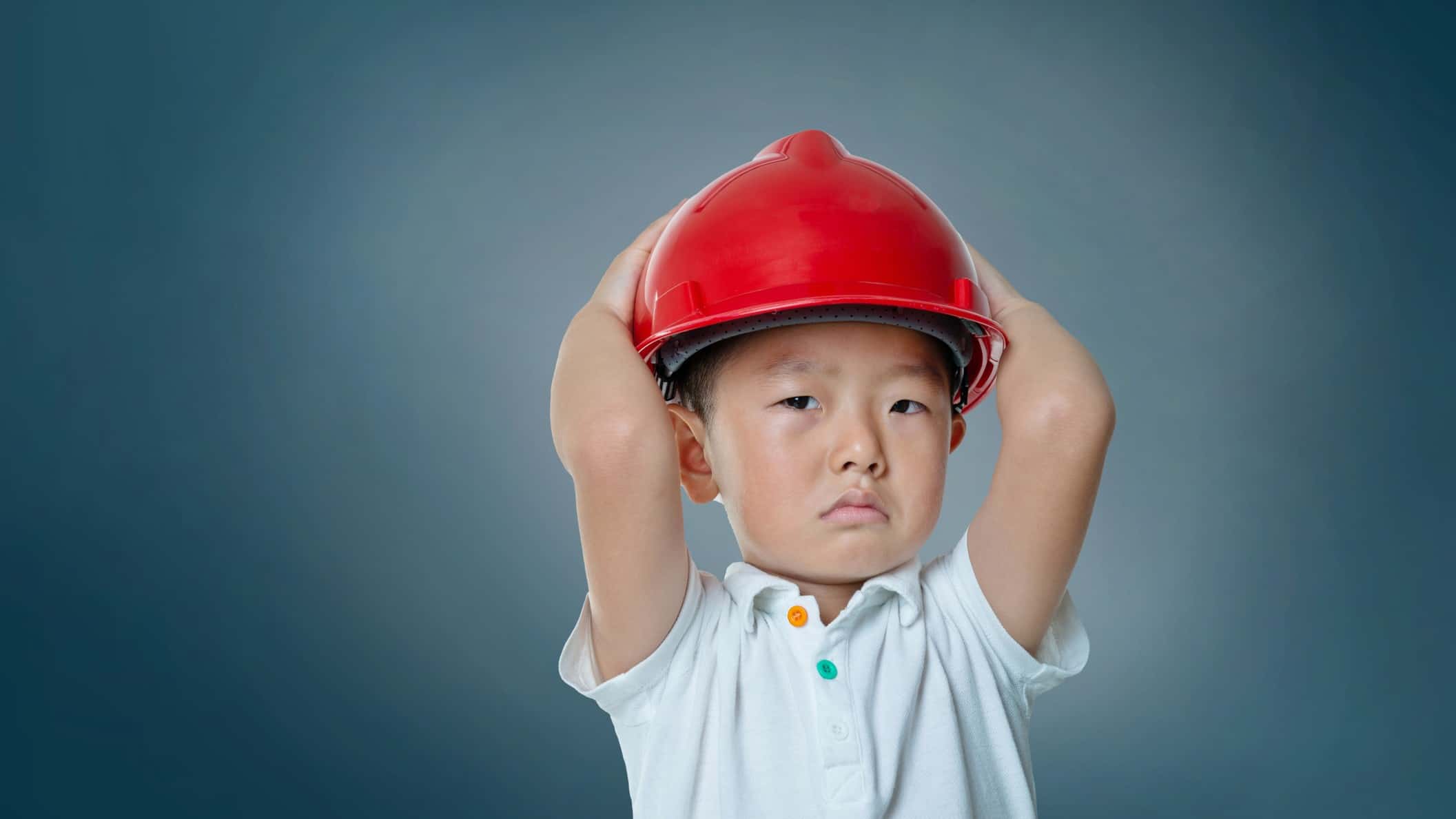 Young boy wearing a red hard hat frowning with his hands on his head.