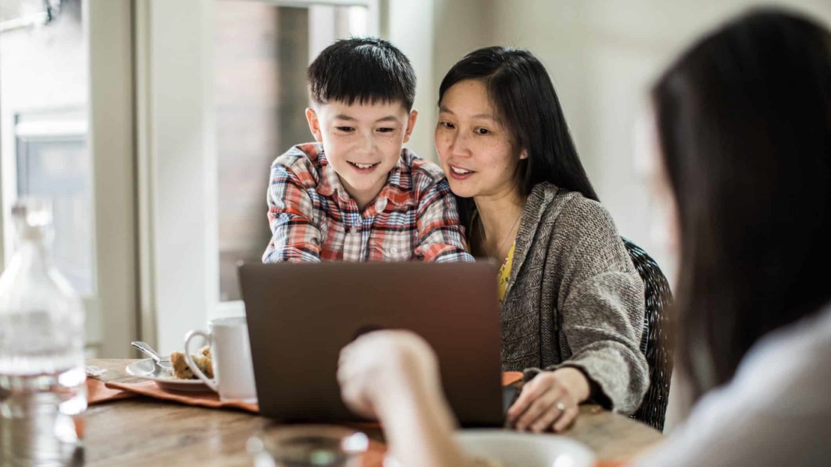 A mother helping her son use a laptop at the family dining table.