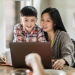 A mother helping her son use a laptop at the family dining table.