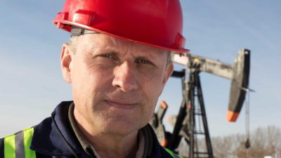 Close up of a miner wearing a hard hat with a solemn look on his face, with an oil drill in the background.