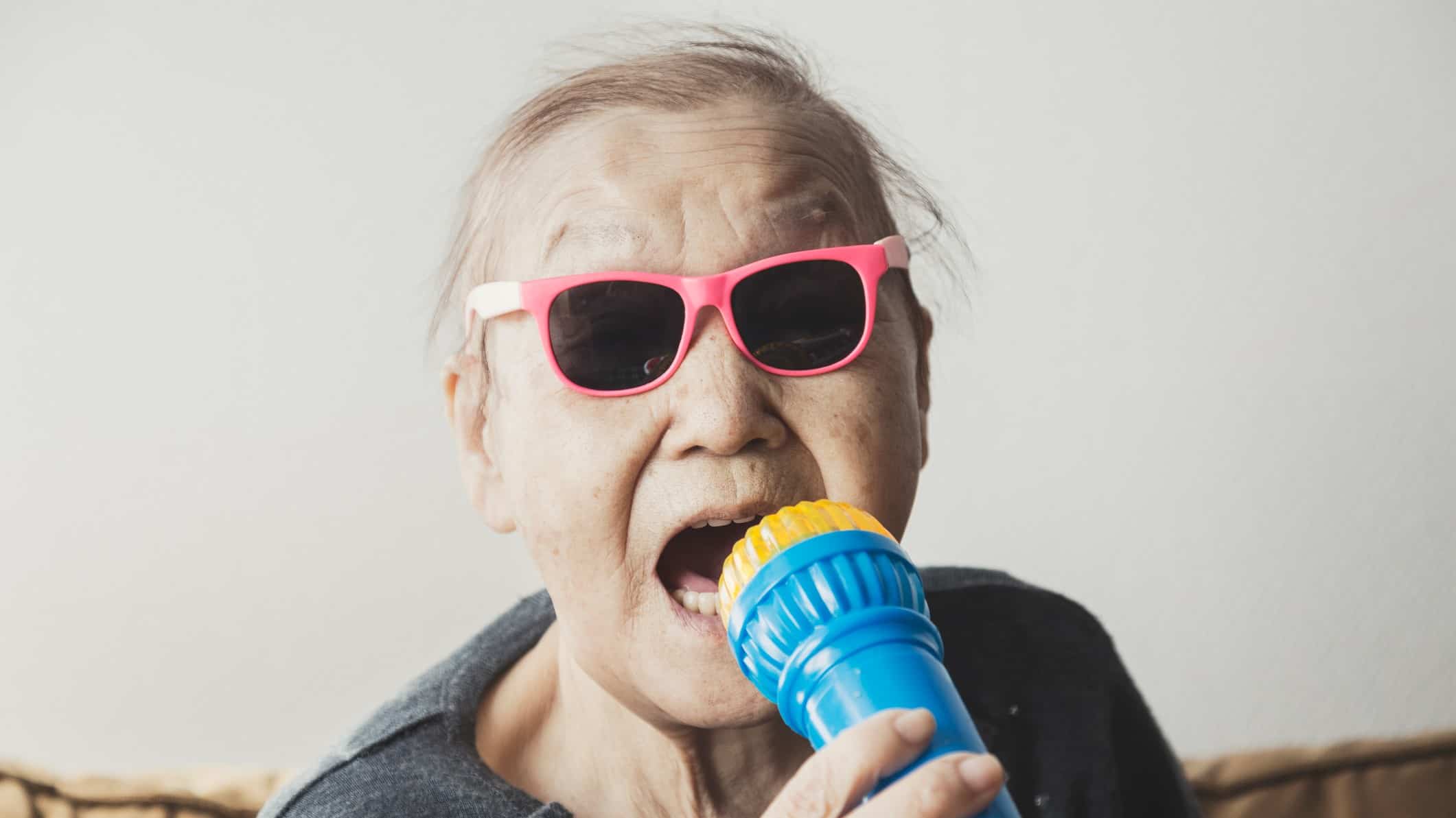 An elderly Asian man sings karaoke into a microphone, wearing cool pink sunglasses.