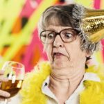 An older woman wearing a wonky party hat looks unpleasantly at a glass of wine in her hand.