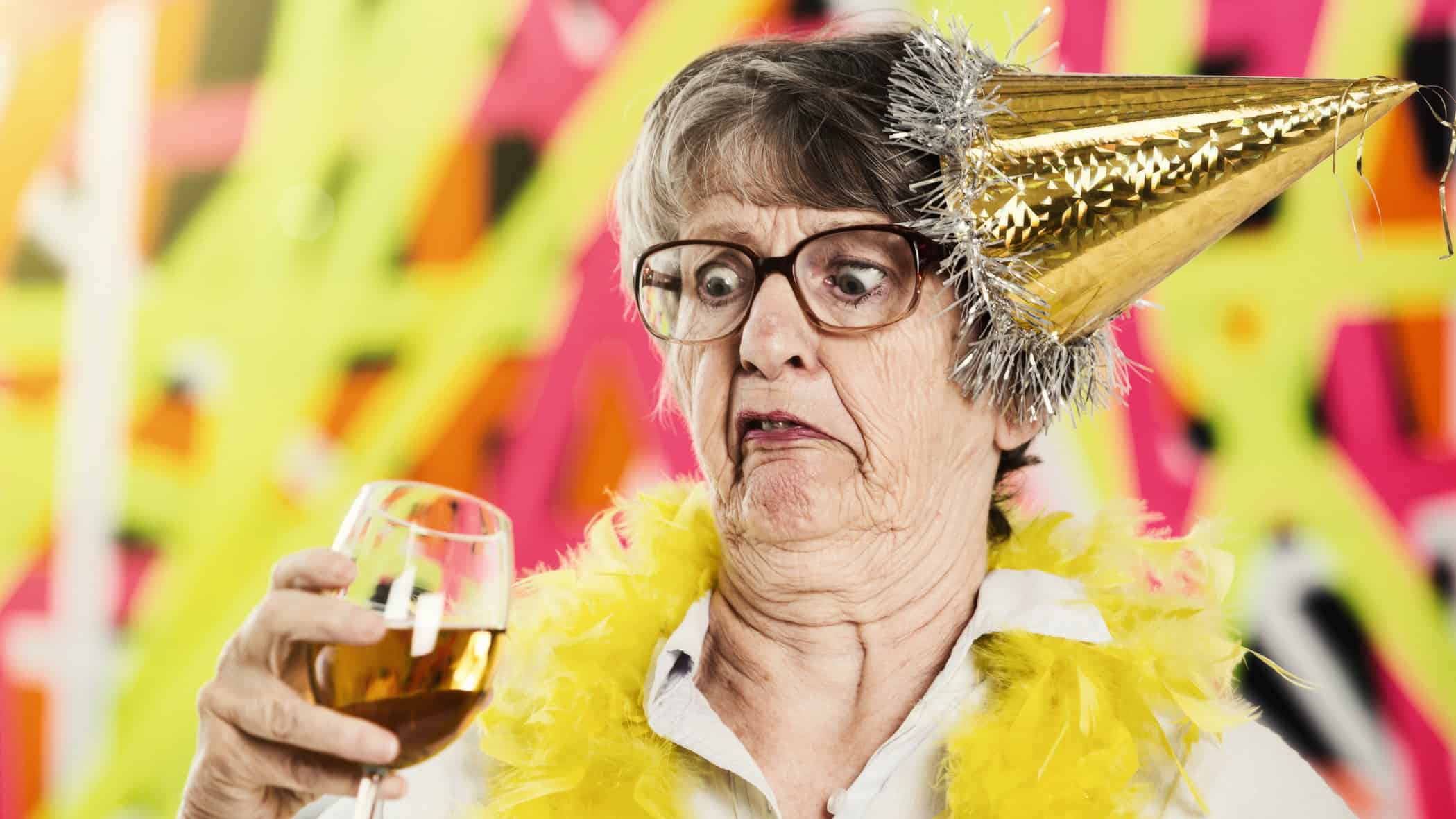 An older woman wearing a wonky party hat looks unpleasantly at a glass of wine in her hand.