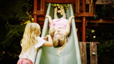 A young girl stands by the slide in a playground while her friend slides down head first and on her back.