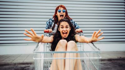 One twenty-something girl pushes her friend in a trolley directly towards the camera, both very excited.