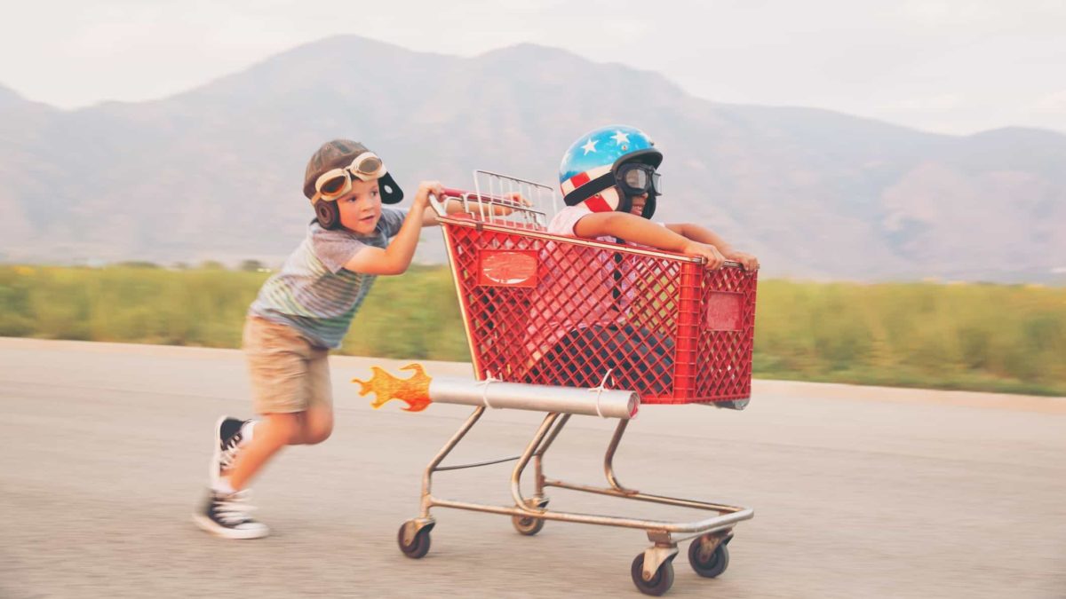 A young boy pushing his friend in a shopping trolley race along the road.