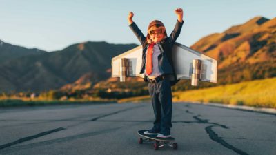 Young boy in a suit and red tie standing on a skateboard with a rocket on his back, arms in the air showing confidence.