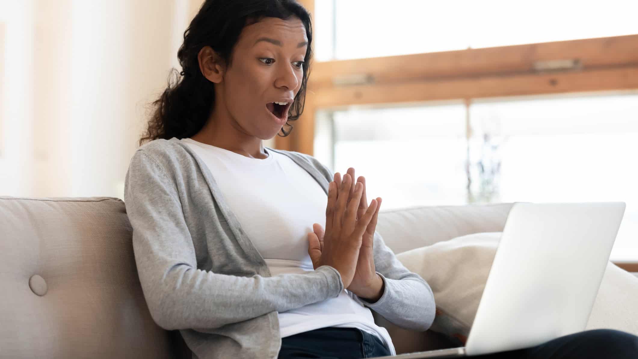 Young woman sitting on nice furniture is pleasantly surprised at what she's seeing on her laptop screen.