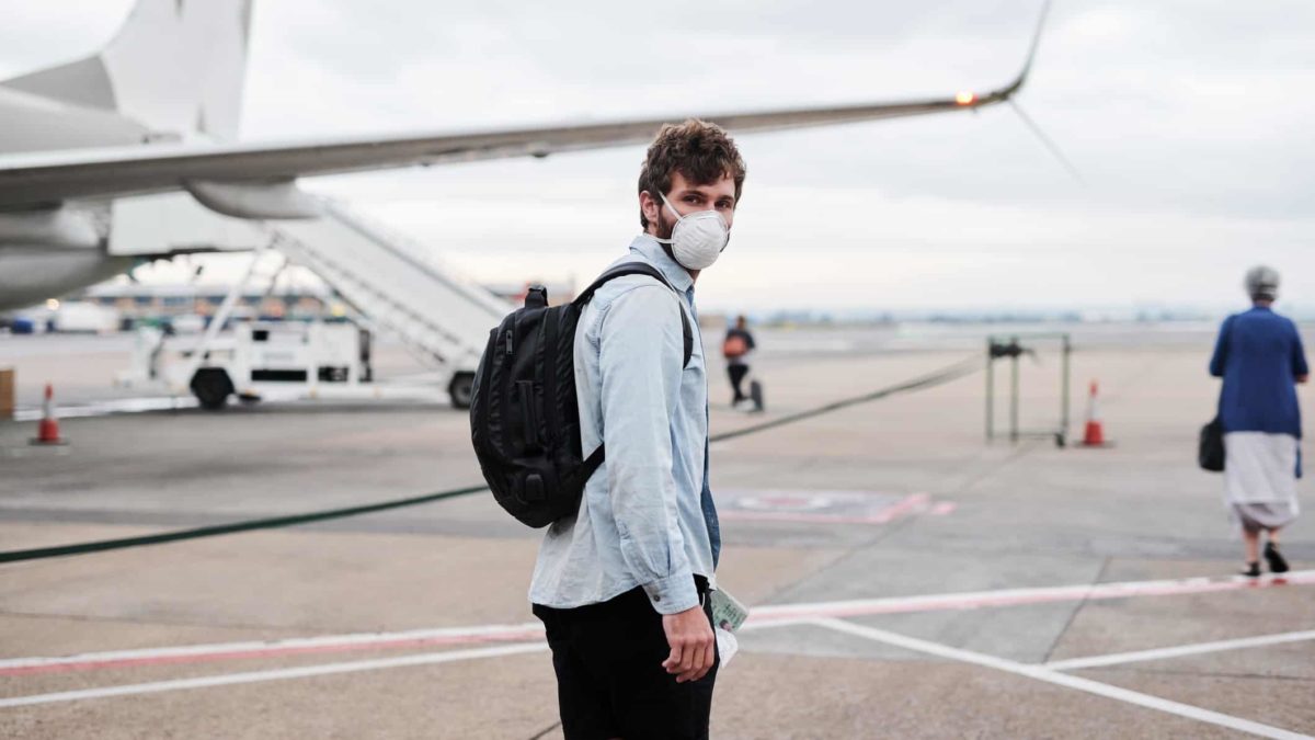 A younger man wearing a face mask turns to look at the camera as he walks towards a plane on the tarmac.