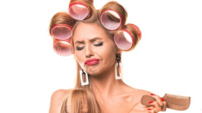 Woman with large rollers in her hair and using cosmetics looking frustrated down at the ends of her hair.