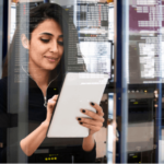 Female cyber security expert surrounded by data on glass screens and looking down at a tablet.