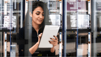 Female cyber security expert surrounded by data on glass screens and looking down at a tablet.