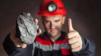 A coal miner wearing a red hard hat holds a piece of coal up and gives the thumbs up sign in his other hand